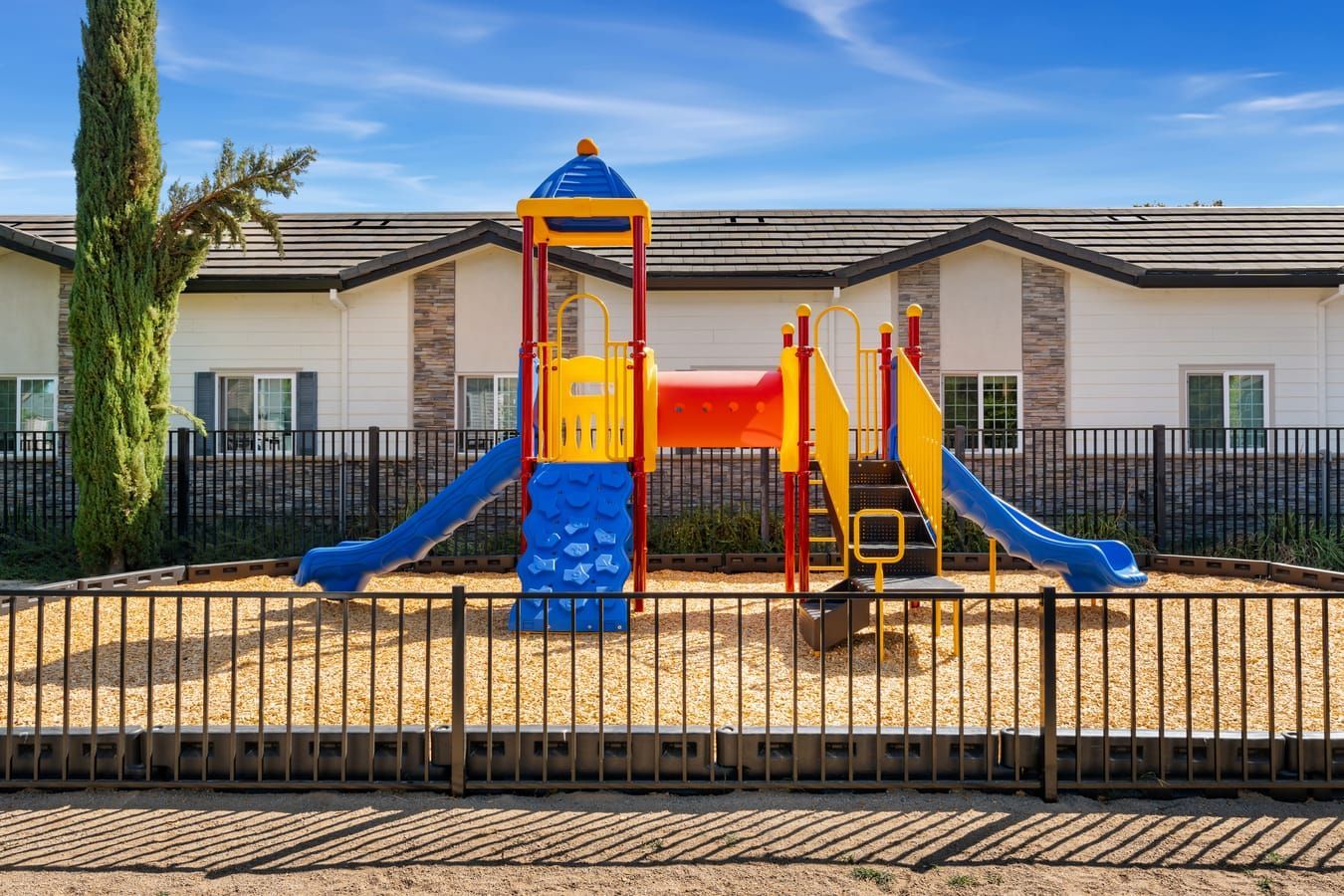 Colorful playground with blue and yellow slides behind a black fence at a residential building