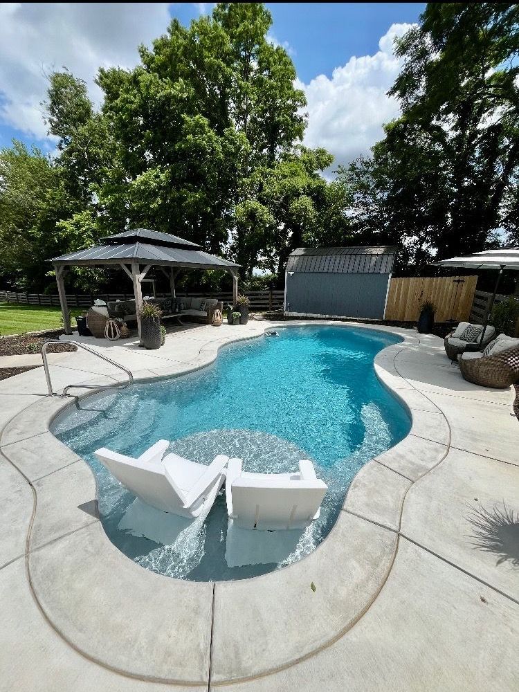 Swimming pool with two white lounge chairs, a gazebo, and blue water. Sunny day with trees.