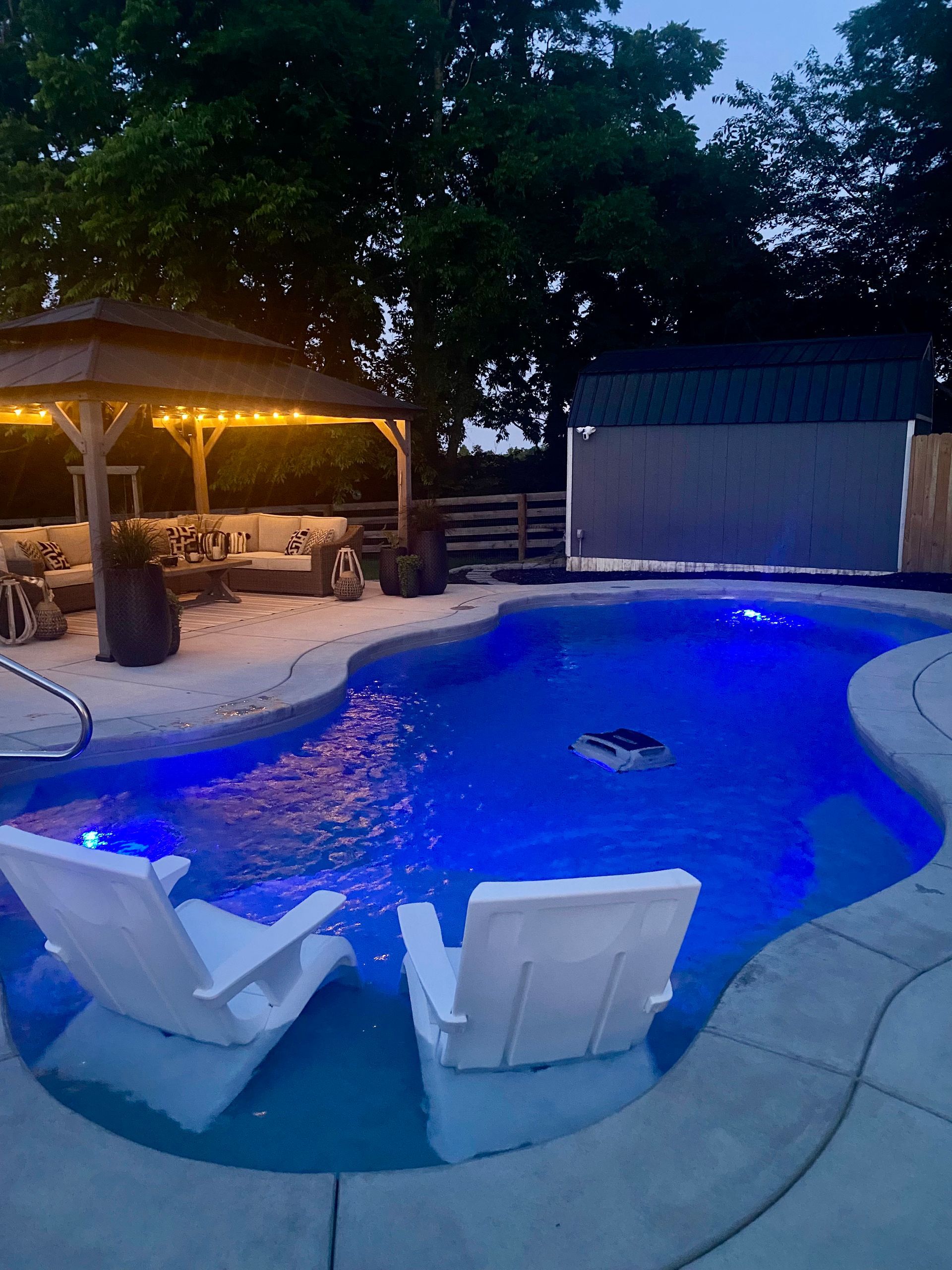 Nighttime view of a backyard pool with blue lights, lounge chairs, and a gazebo with string lights.