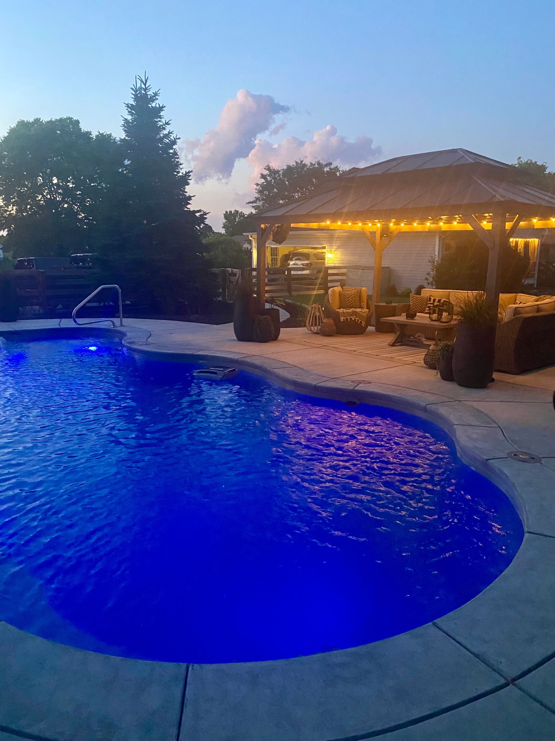Blue-lit pool at dusk with gazebo strung with lights, trees in background.