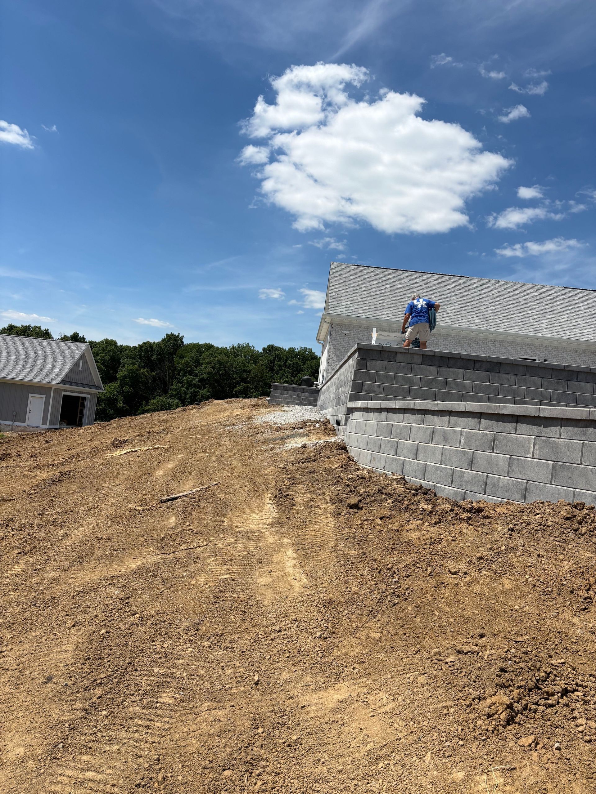 Worker on a cinder block house roof beside a dirt slope under a bright blue sky