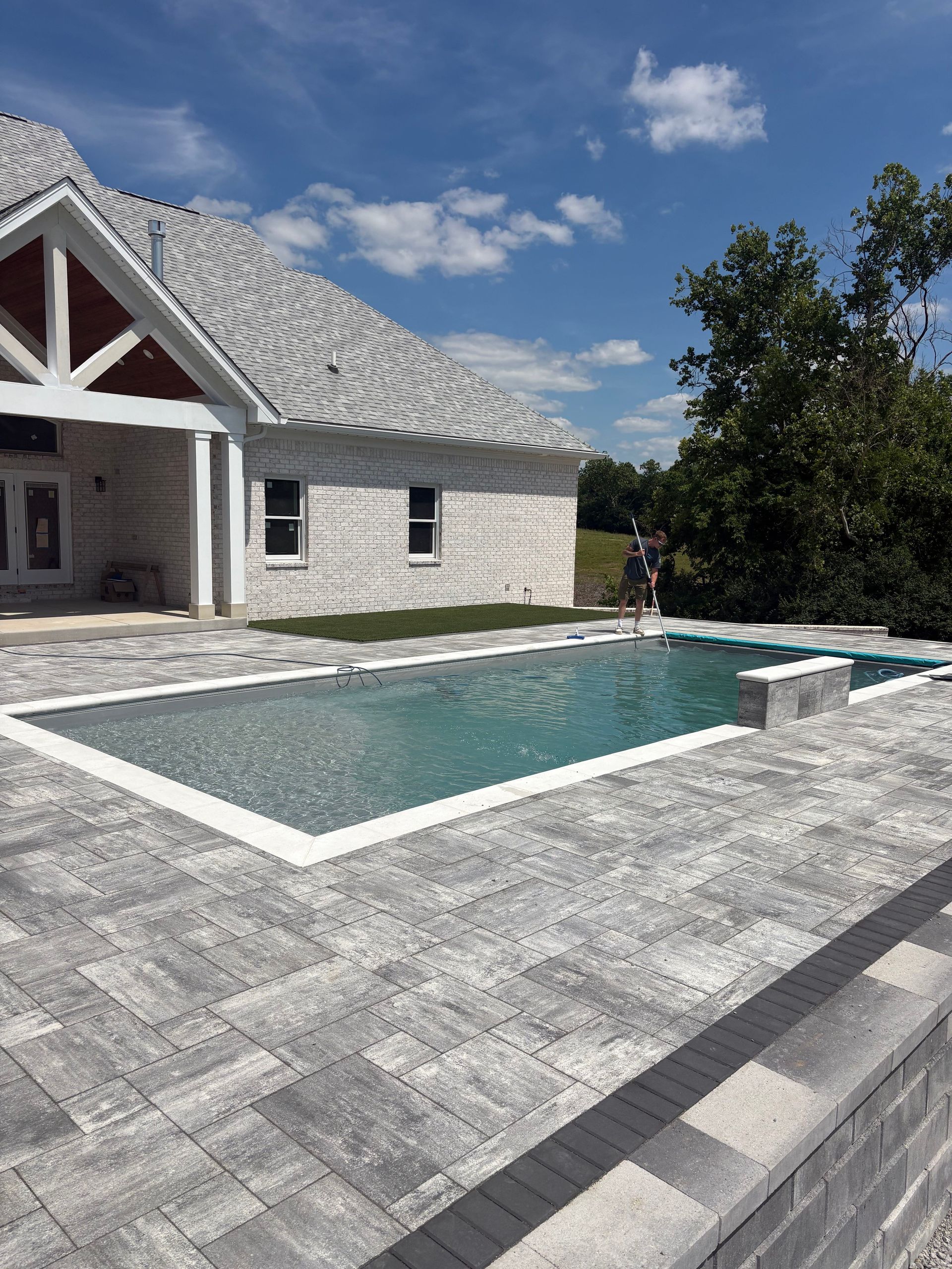 Stone patio with a rectangular pool beside a house under a partly cloudy sky
