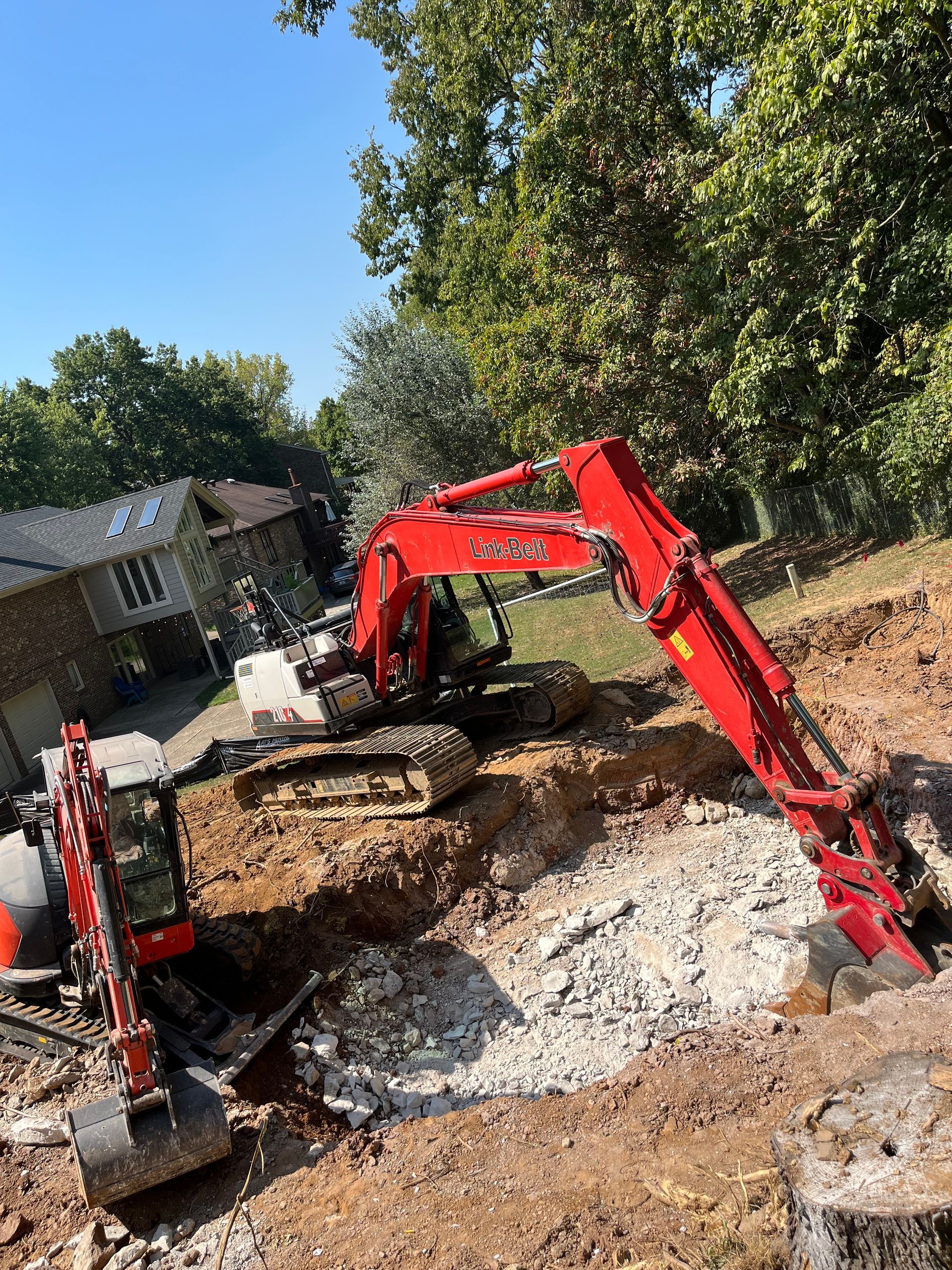 Red excavator digging dirt at a construction site beside a house and trees