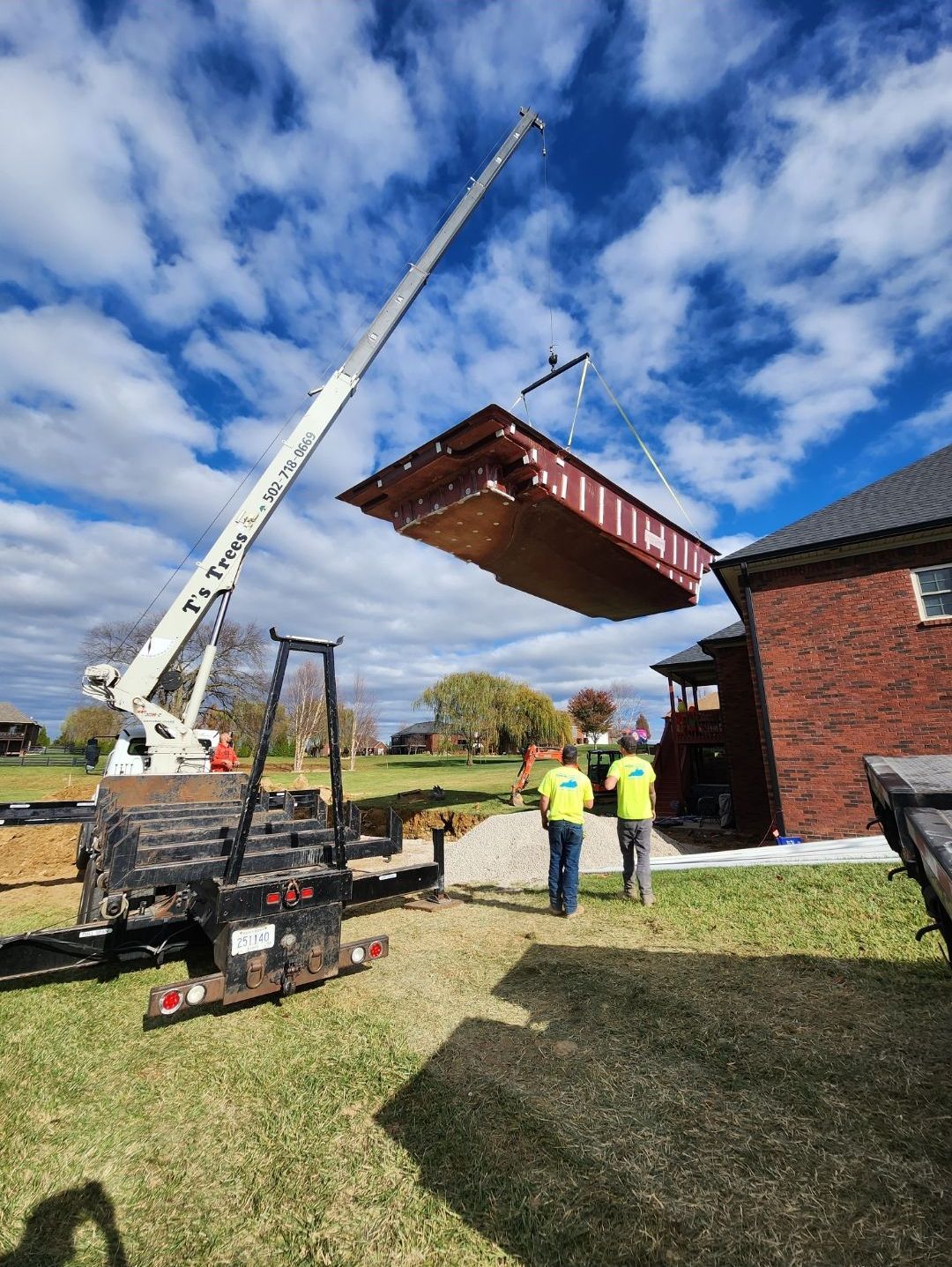A crane lifting a large rectangular structure over a house with two workers nearby under a cloudy sky.
