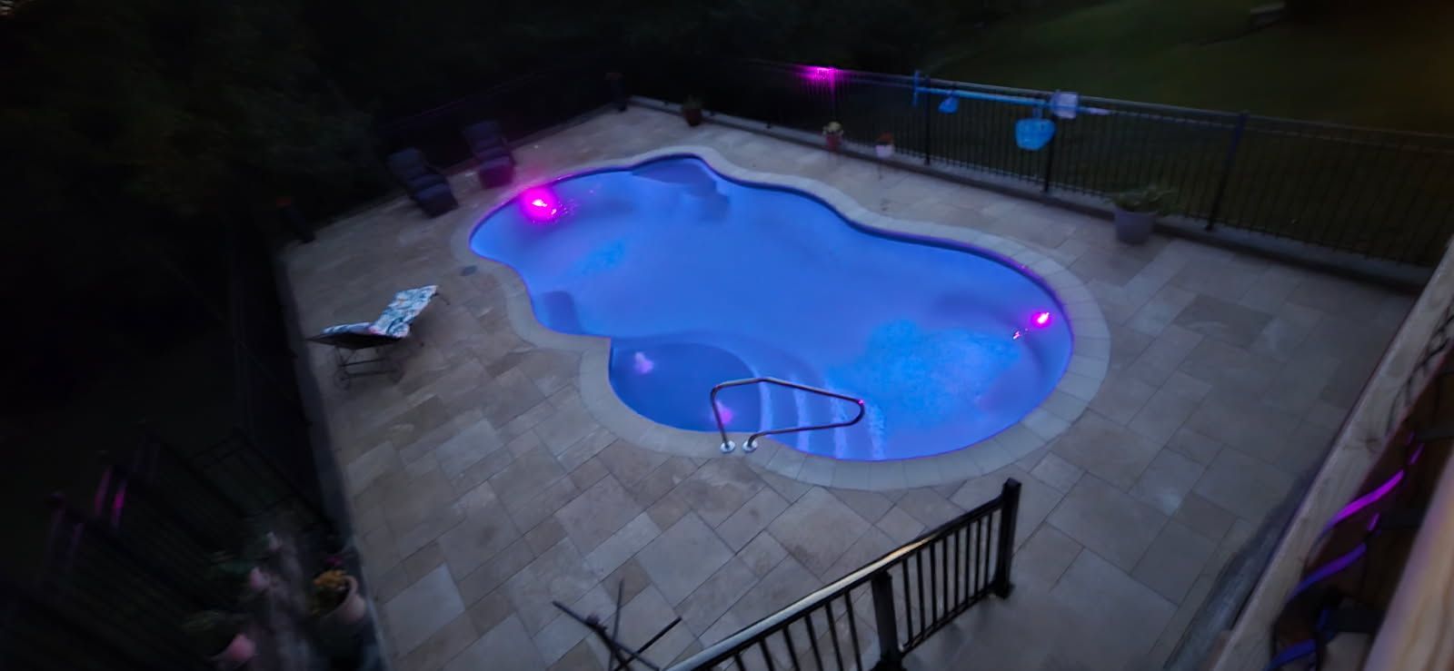 A nighttime view of a pool with blue water and pink lights, surrounded by a patio.