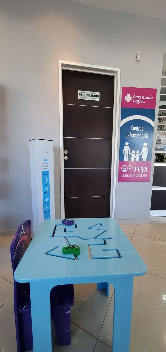 Vestíbulo con una pequeña mesa azul, cordones para credenciales, una puerta y carteles informativos en la pared.