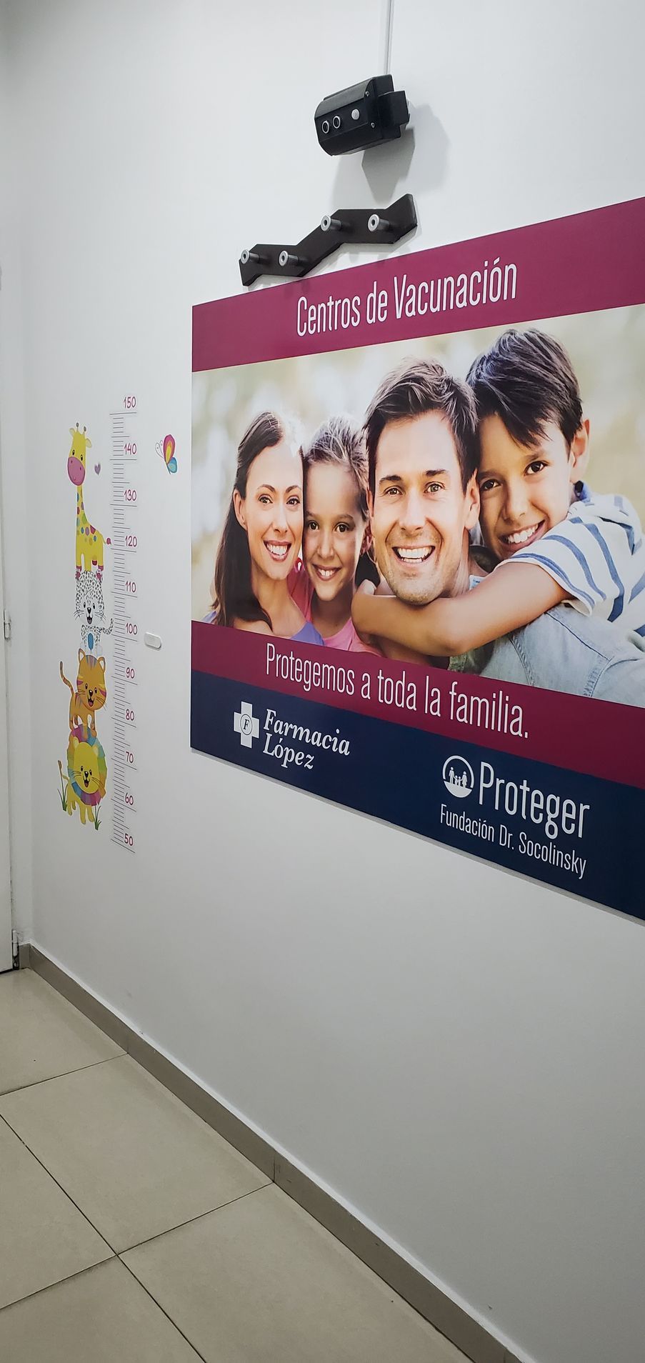 Póster en la pared del pasillo con una foto de grupo sonriente y un encabezado rosa debajo de una cámara de seguridad.