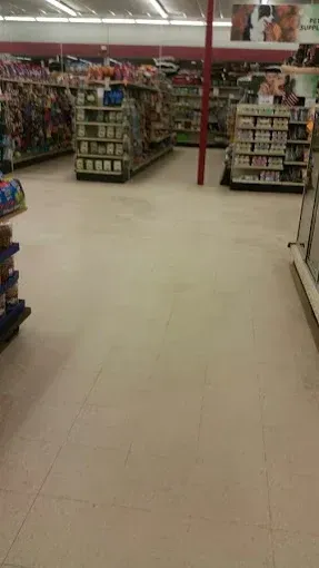 Interior of a store with shelves stocked with merchandise, beige tiled floor, and a red pole in the middle.