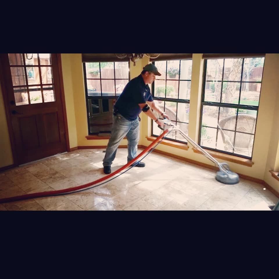Man cleaning a tiled floor with a rotary machine in a room with large windows and a wooden door.