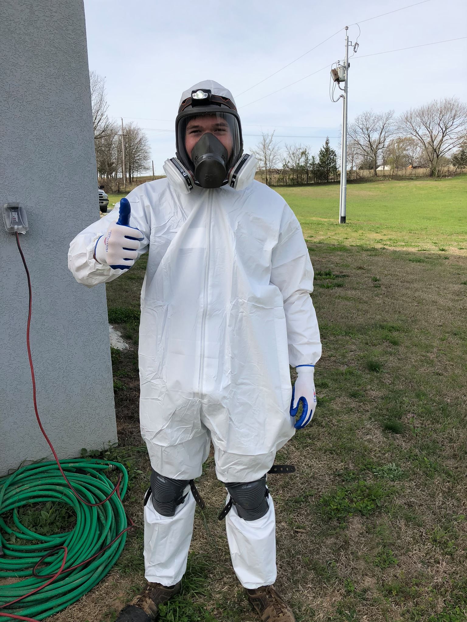 Man in white hazmat suit and respirator gives thumbs up outside.