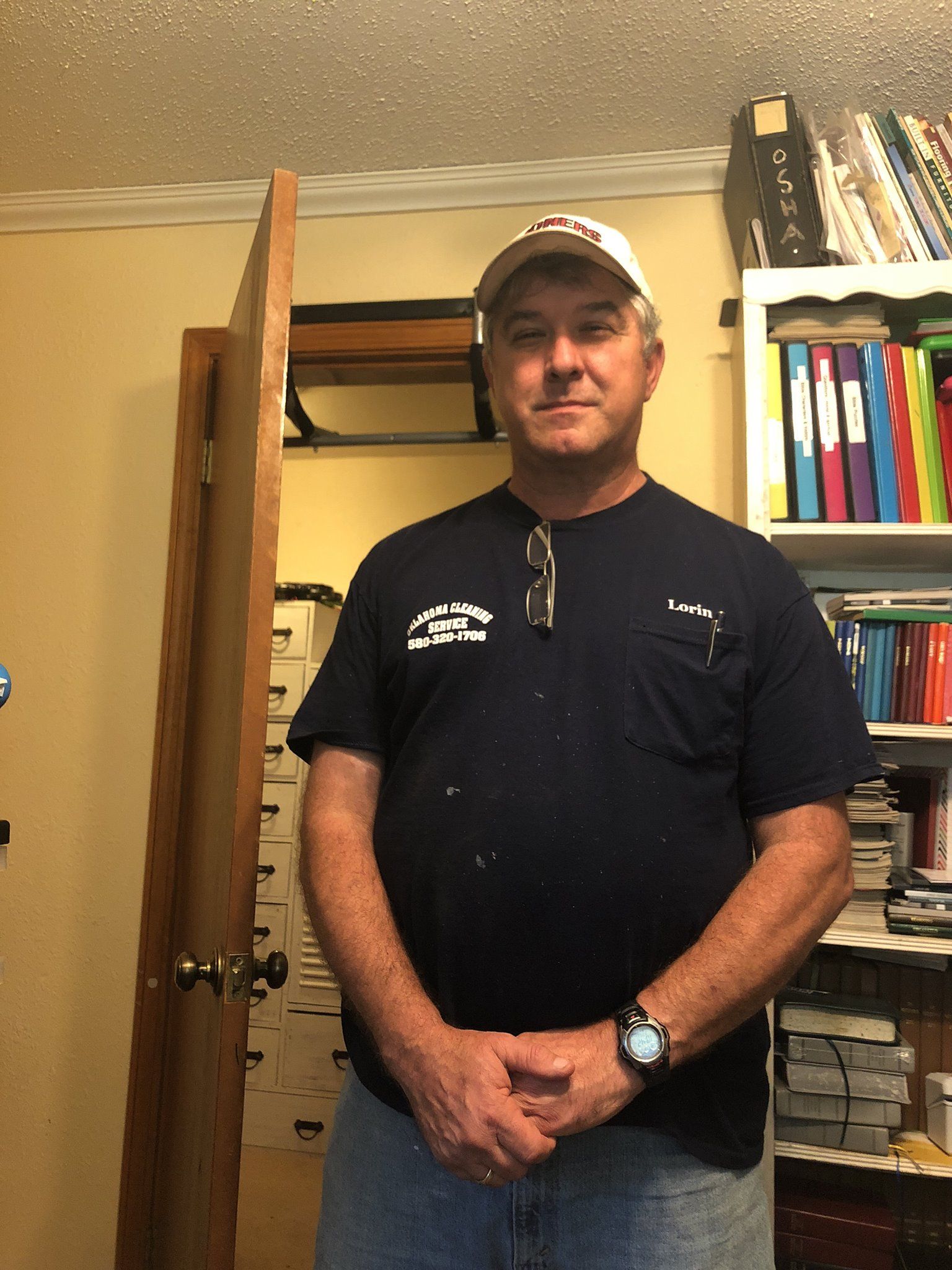 Man with a cap and t-shirt stands in a cluttered office; arms crossed.
