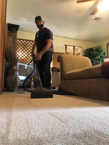 Man in mask cleans beige carpet with machine in living room.