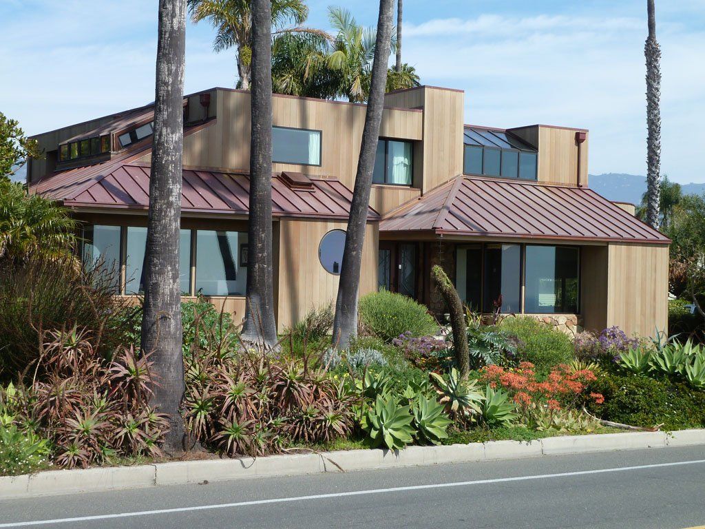 Modern house with brown siding and reddish-brown roof, surrounded by lush landscaping and palm trees.