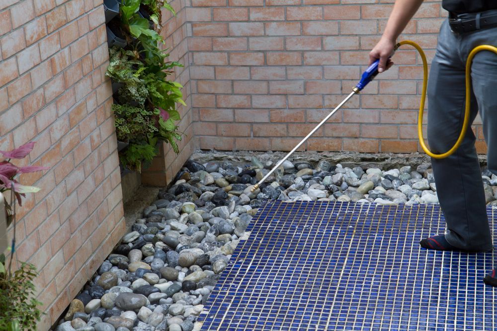 Person spraying a patio with a wand, next to a rock garden and brick wall — Hancock Pest Control in Mardi, NSW
