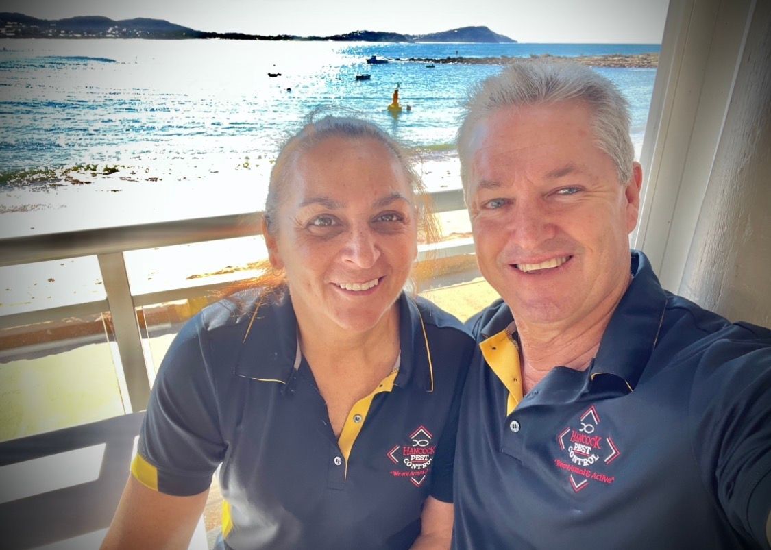 Smiling couple in navy polos taking a selfie on a boat with bright water and shoreline behind them — Hancock Pest Control in Mardi, NSW