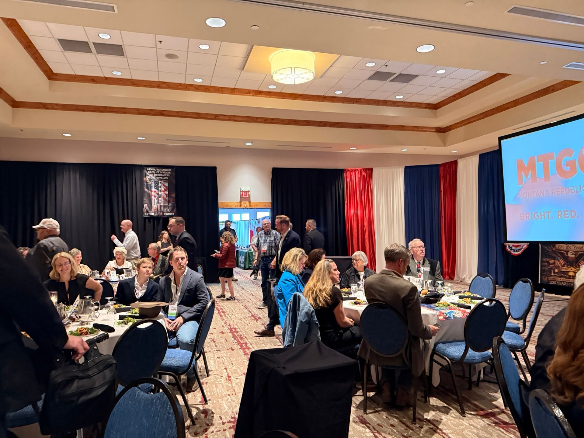 People seated at round tables in a banquet hall during a conference or event, with a stage and “MTG” screen.