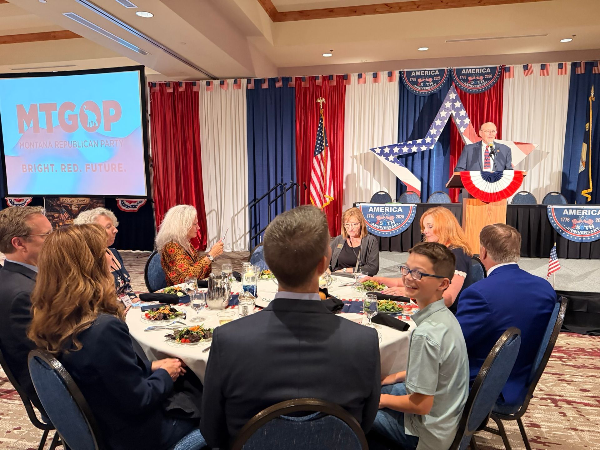 Audience watches a speaker at a red, white, and blue political event with an MTGOP screen.