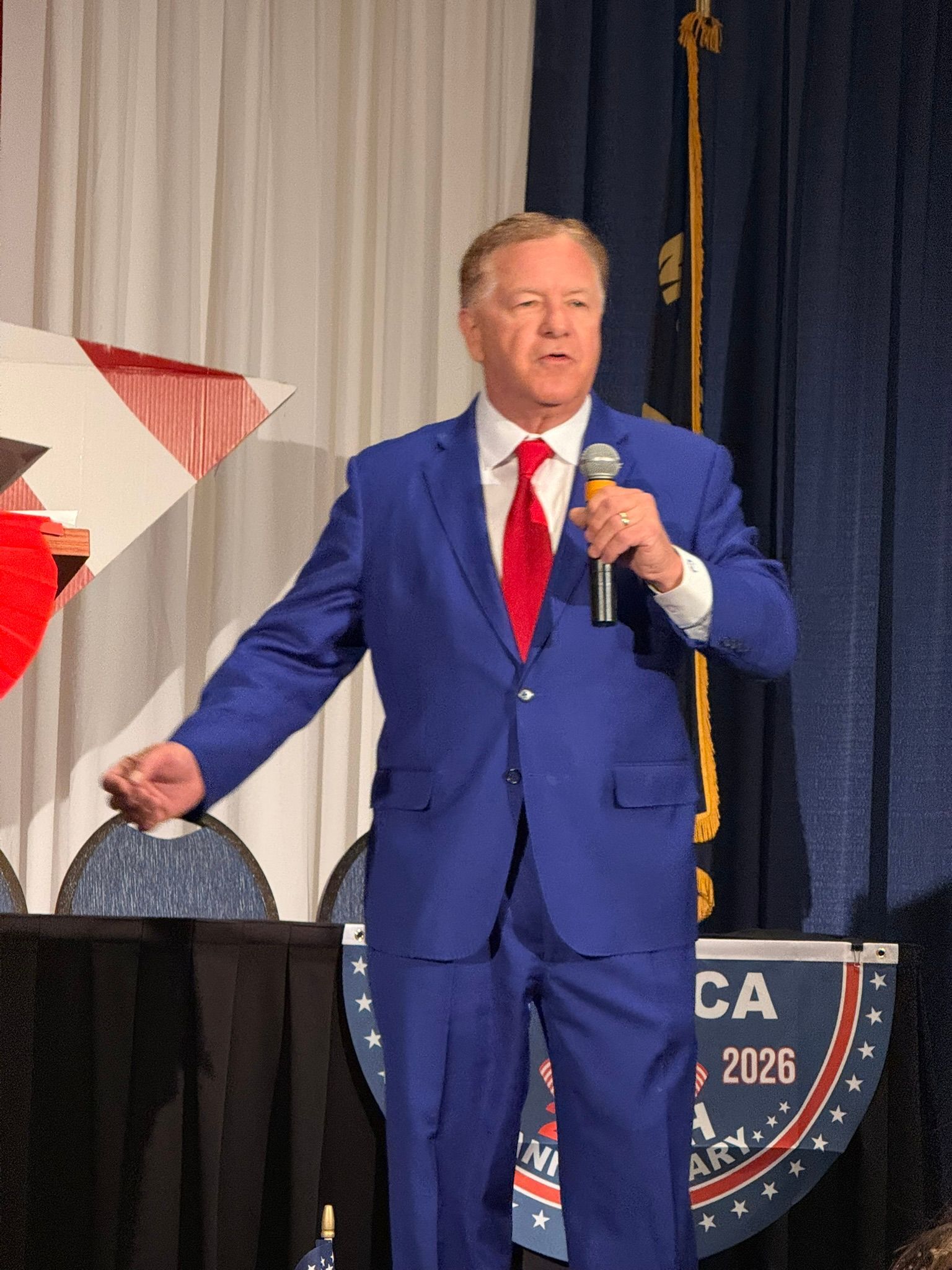 Man in blue suit speaking into a microphone at a podium with a campaign-style backdrop