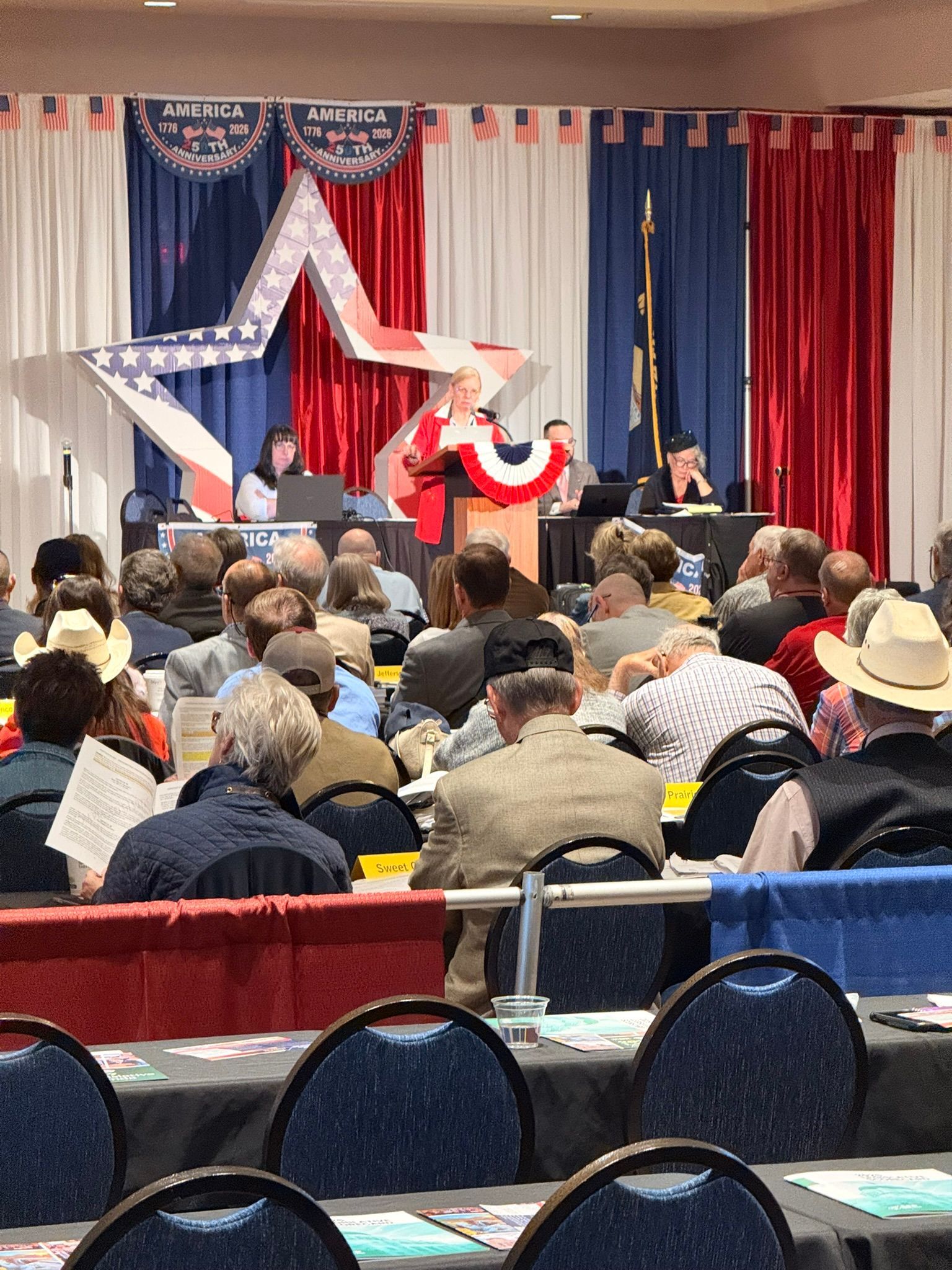 Audience watching a speaker on a patriotic-decorated stage at an indoor event