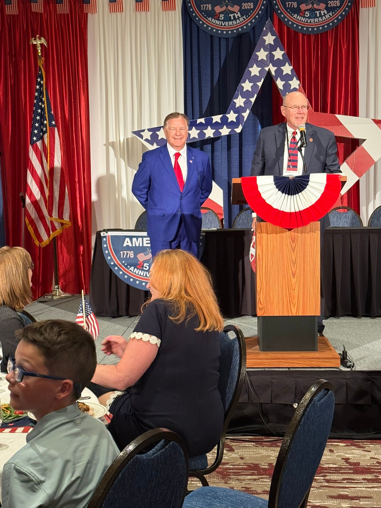 Two men on stage at a patriotic event, one at a podium with red, white, and blue bunting.