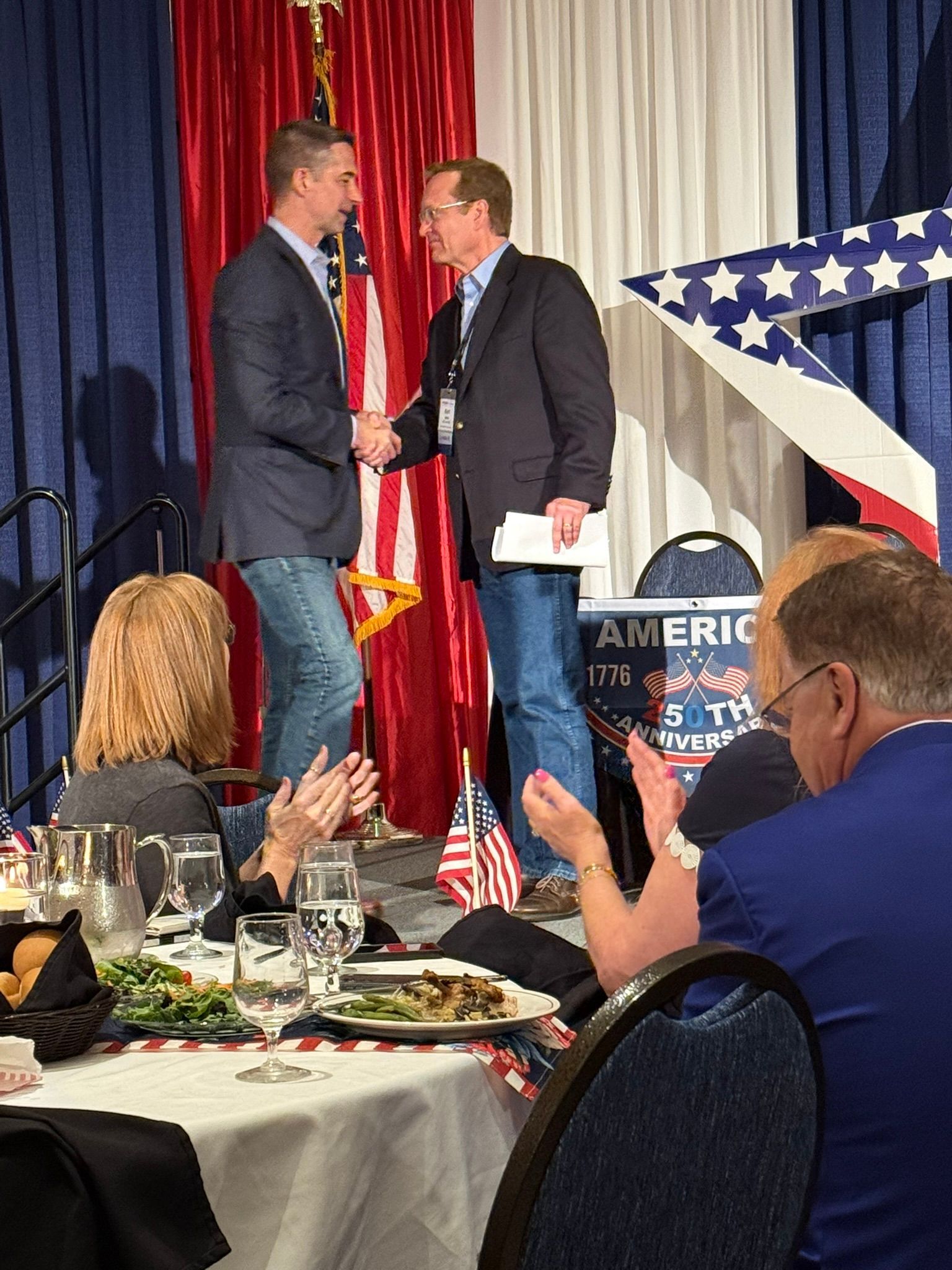 Two men shake hands on a stage at a political event with U.S. flags and an audience clapping.