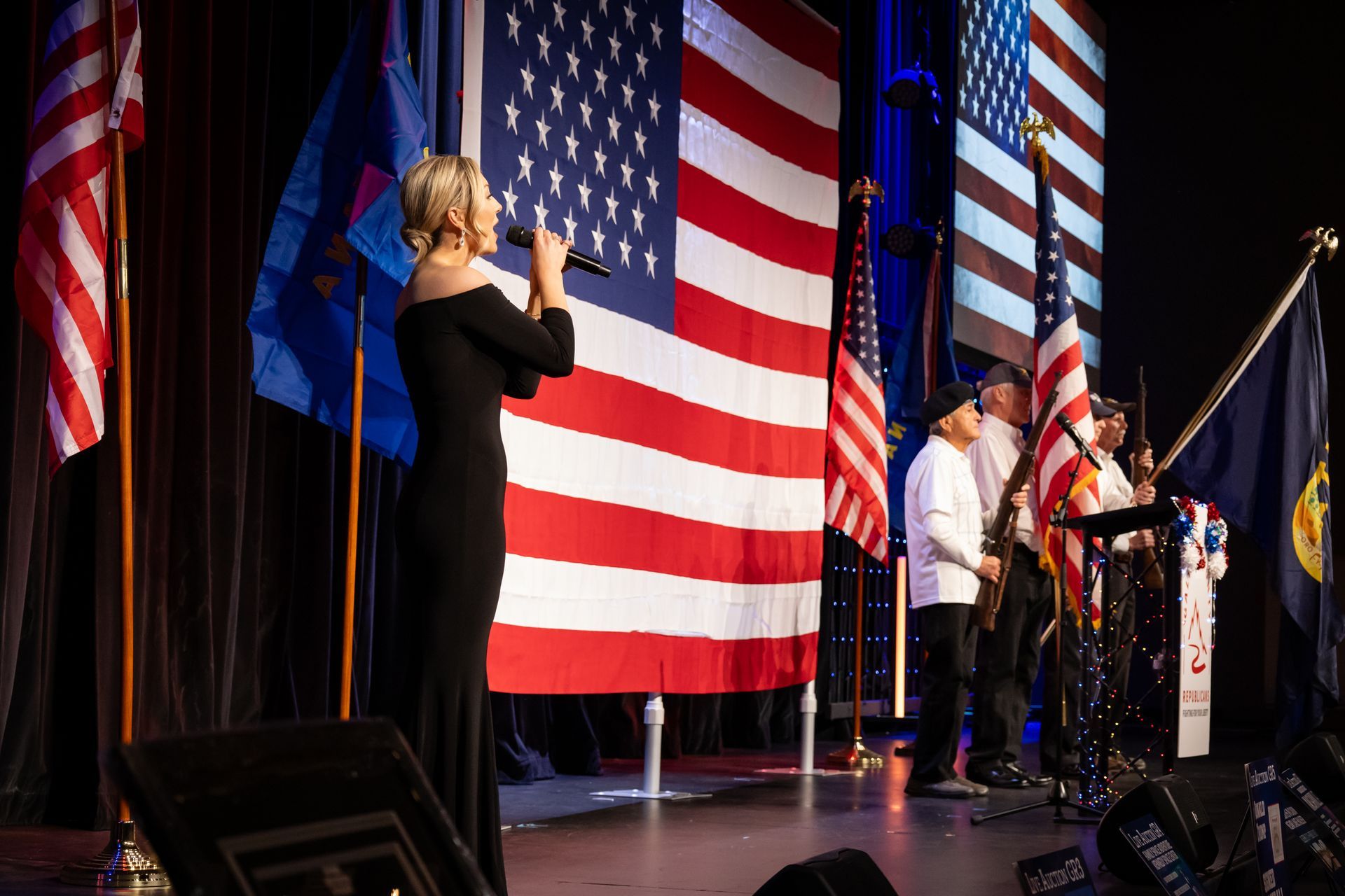 Singer performing on stage before large American flags, with a sign language interpreter nearby.