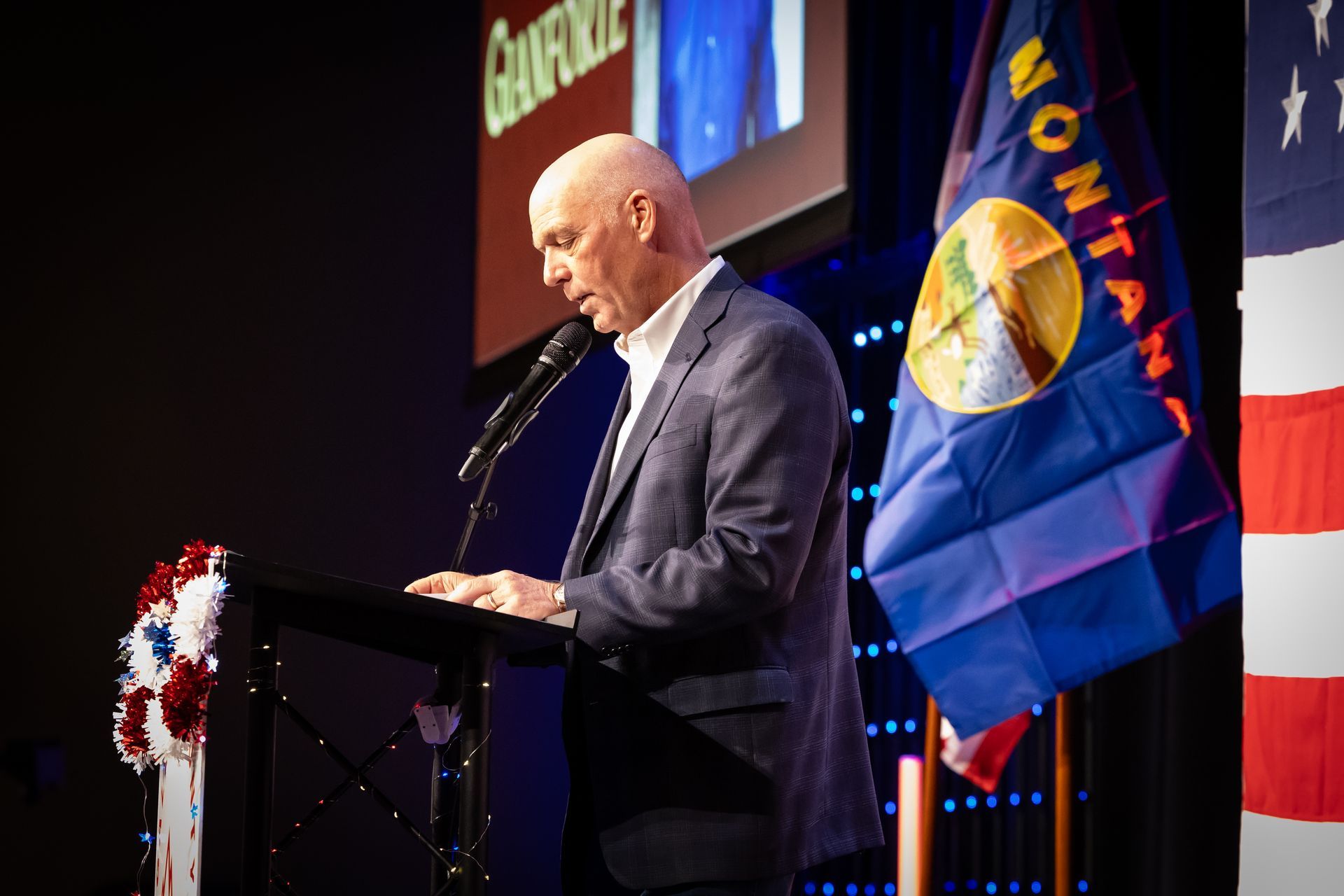 Speaker at podium beside a blue flag on a stage, with a large screen and patriotic decor.