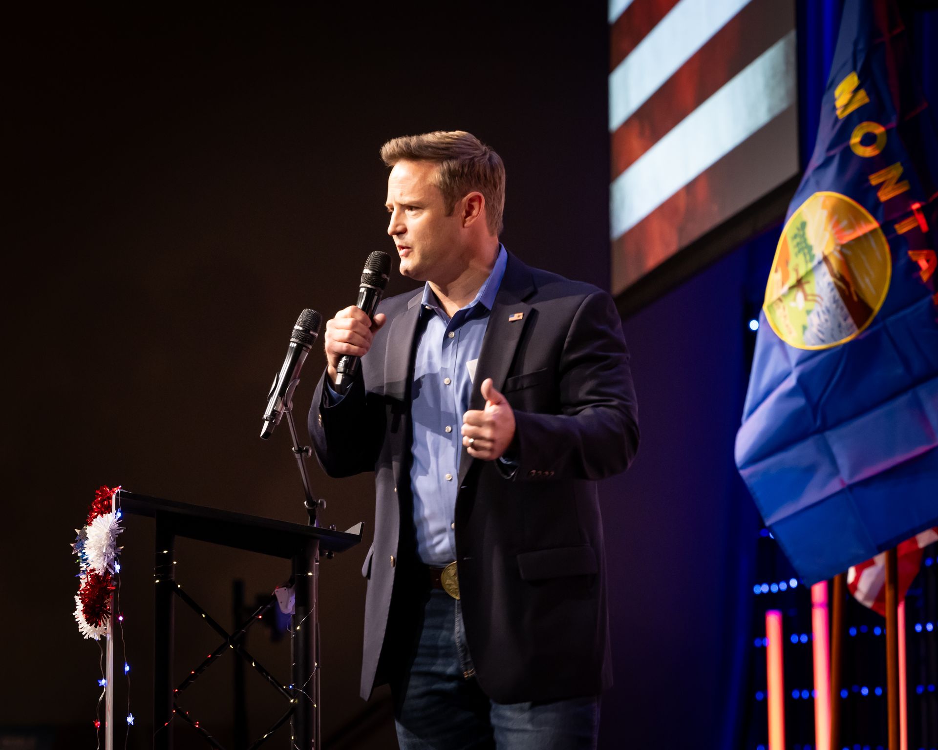 Man speaking into a microphone onstage beside a Montana state flag