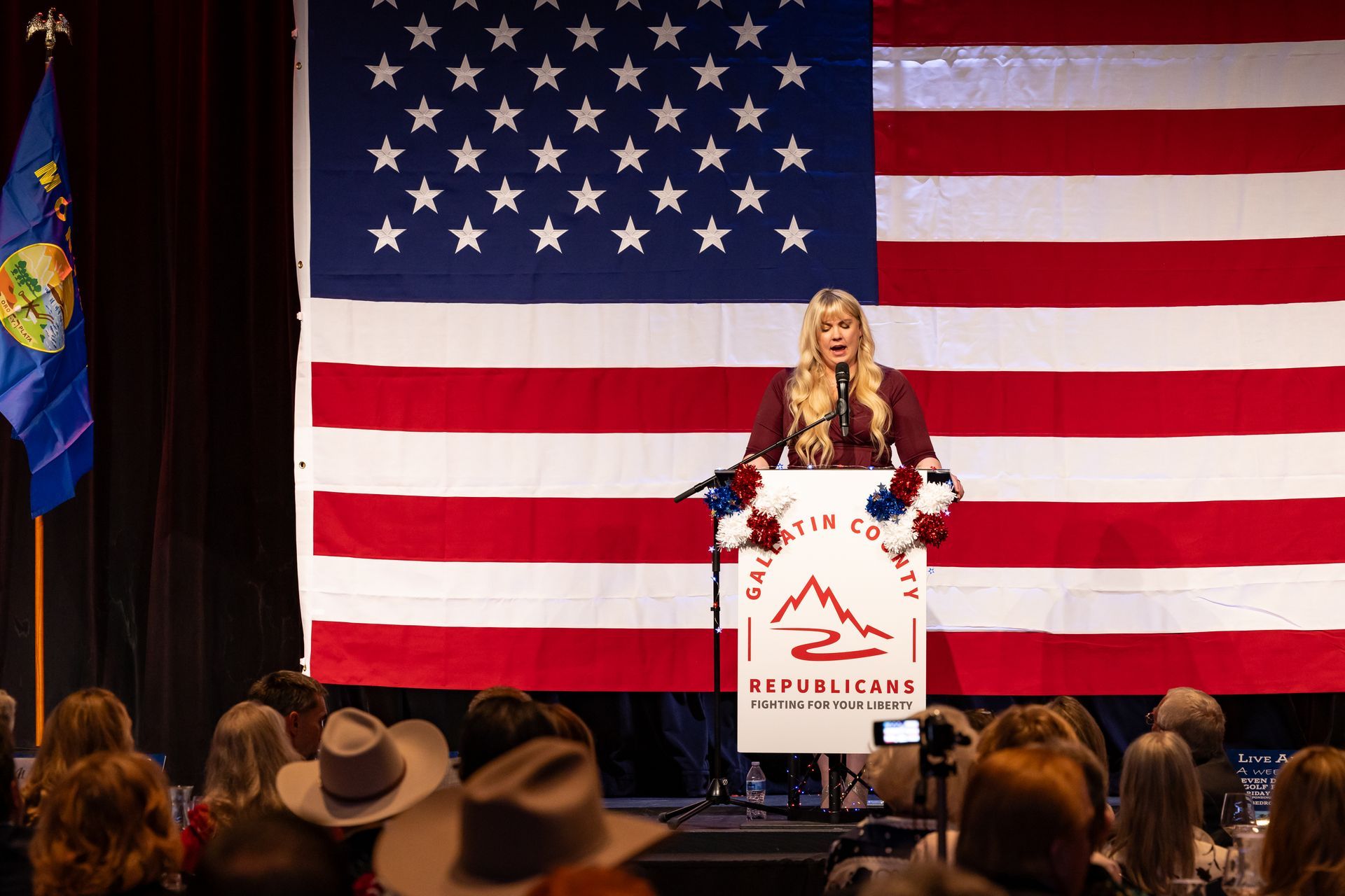 Speaker at podium in front of large American flag, addressing an audience from a stage