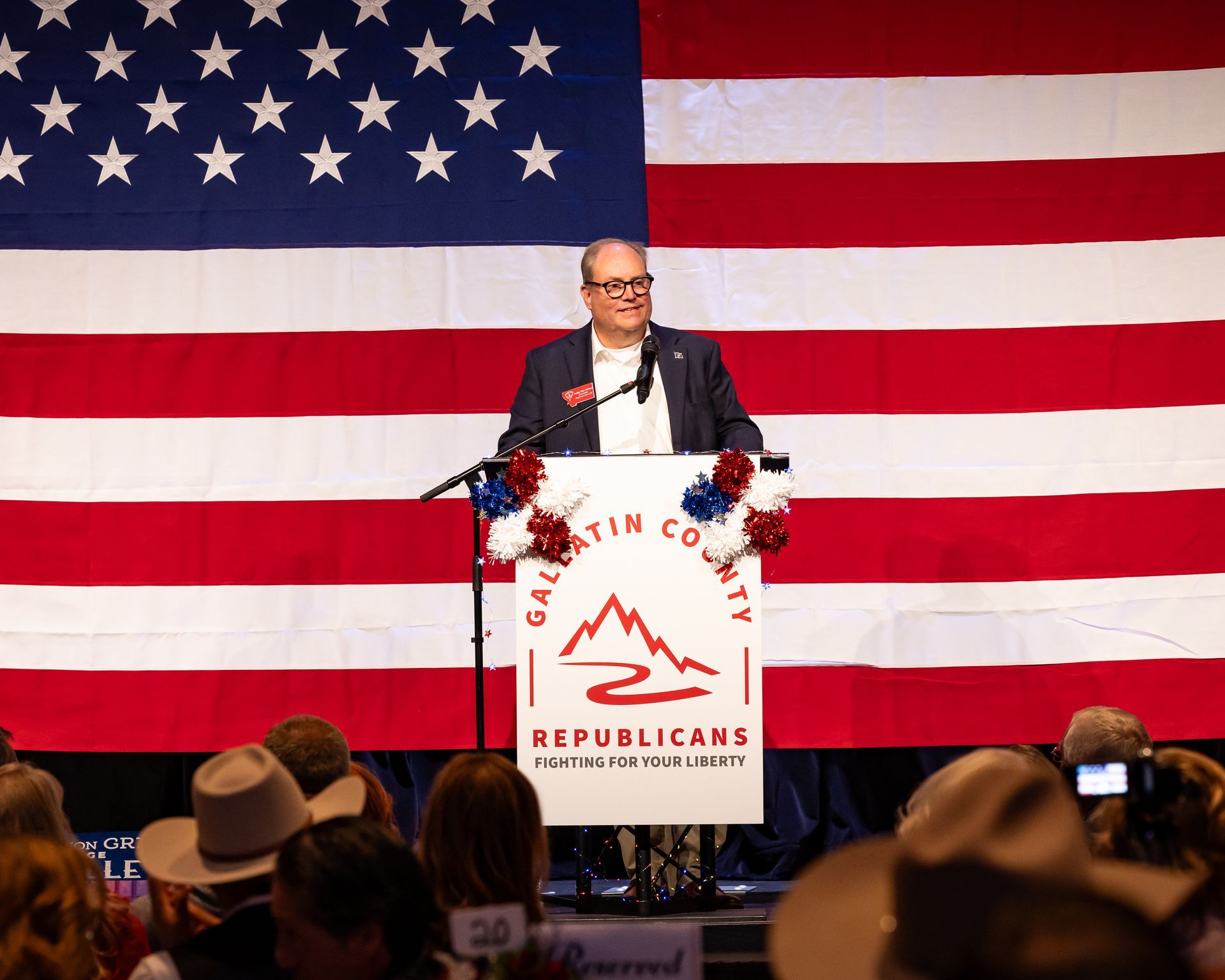 Speaker at podium with Republican Party sign before a large American flag backdrop