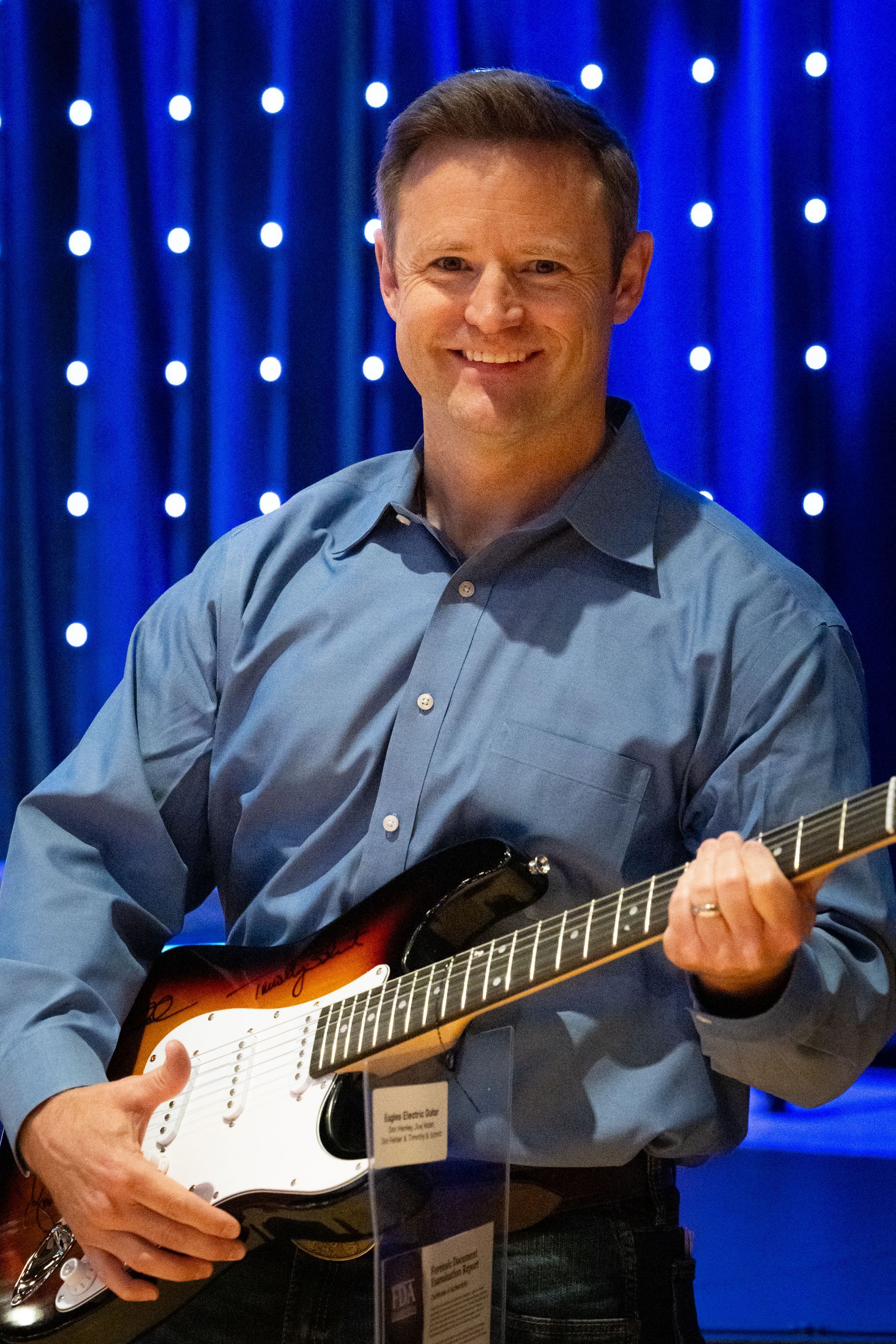 Man smiling while holding an electric guitar on a blue-lit stage