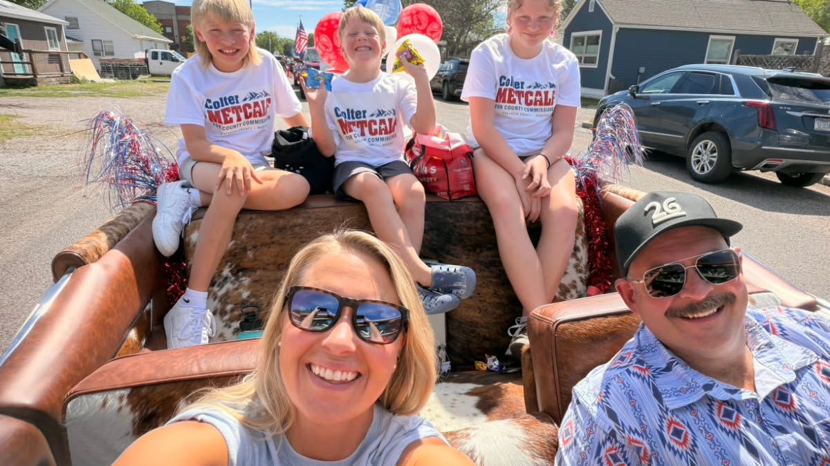 People in sunglasses smile on a wagon ride, with children in red hats behind them at a sunny outdoor event.