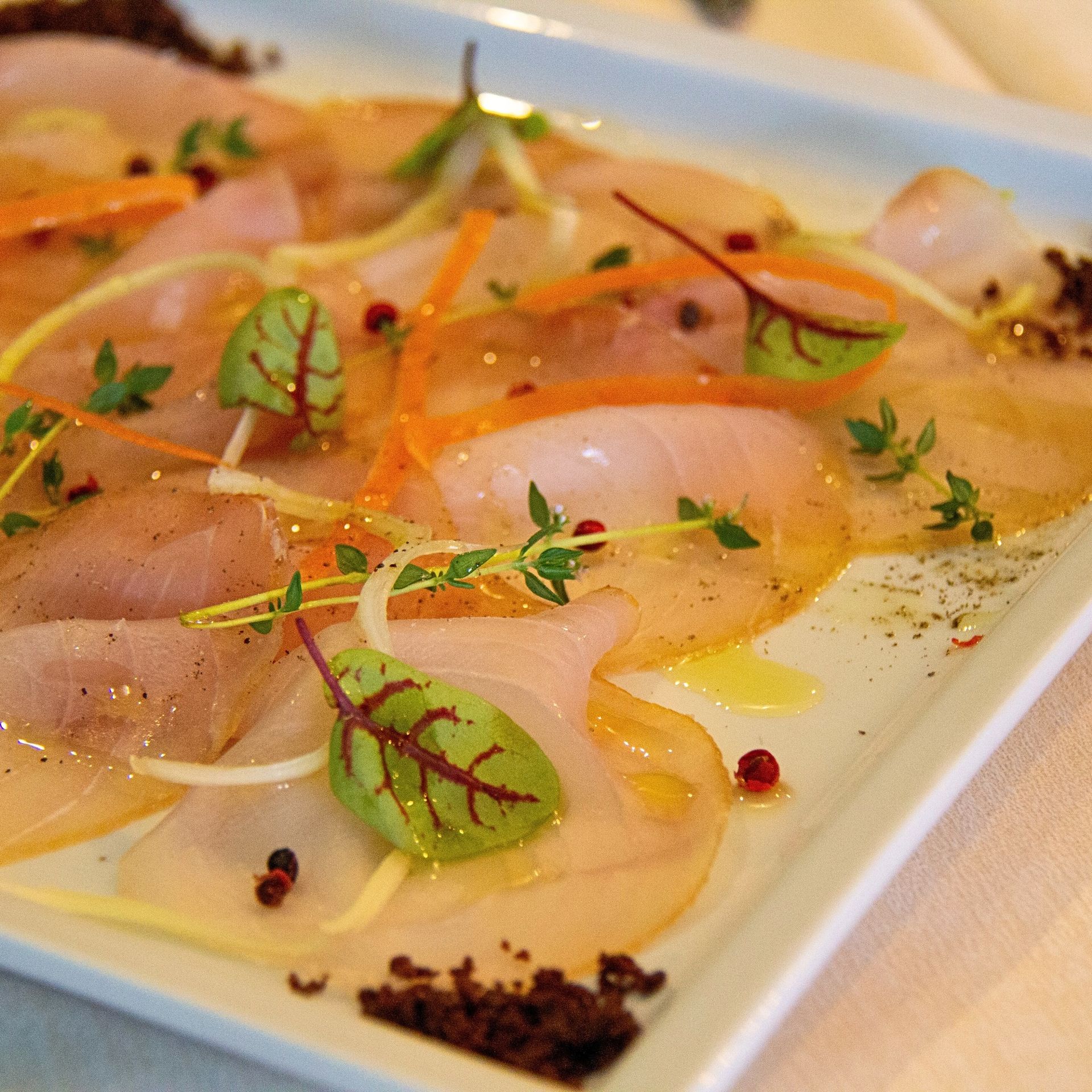Pesce tagliato a fettine sottili, guarnito con erbe aromatiche, agrumi e pepe, servito su un piatto bianco.