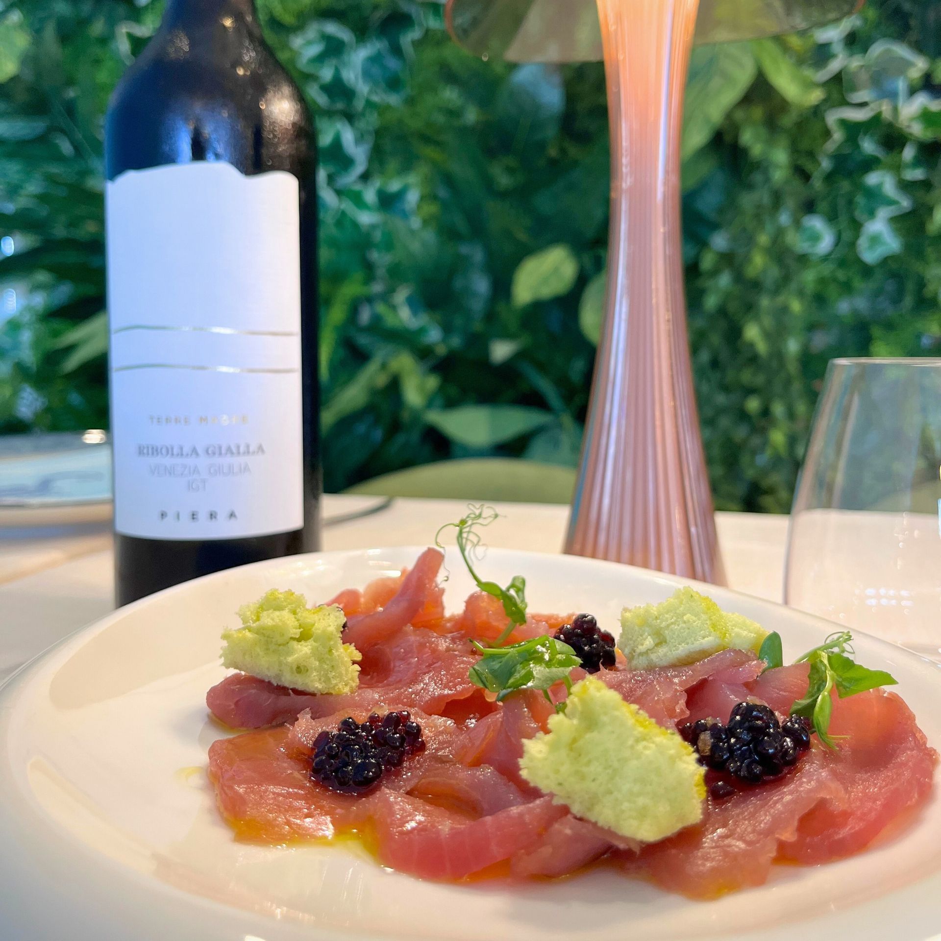 Antipasto di salmone con guarnizione verde e caviale, servito accanto a una bottiglia di vino e un bicchiere su un tavolo.