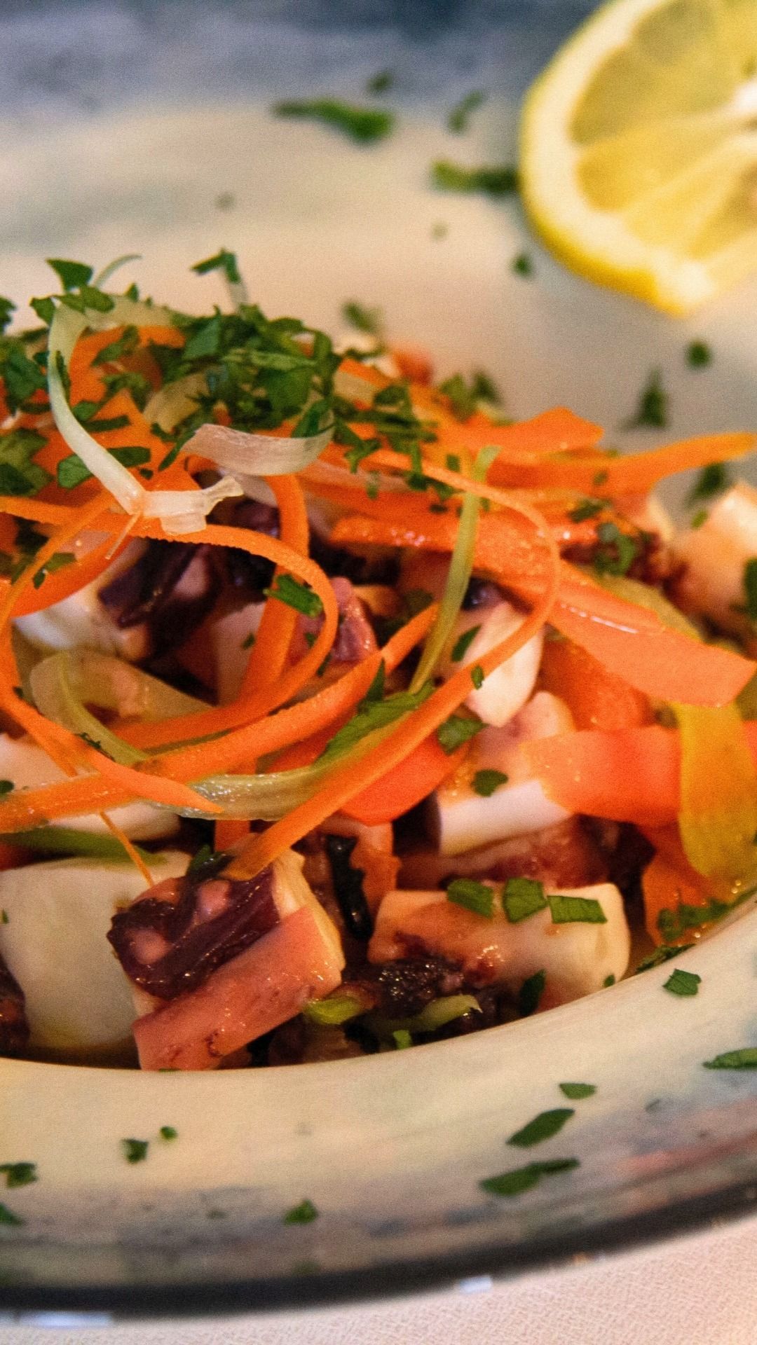 Insalata di frutti di mare colorata, guarnita con striscioline di carota, erbe aromatiche e uno spicchio di limone, servita su un piatto bianco.