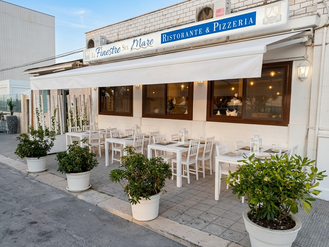 Caffè all'aperto con tavoli e sedie bianchi sotto una tenda a righe, piante in vaso che costeggiano il marciapiede.
