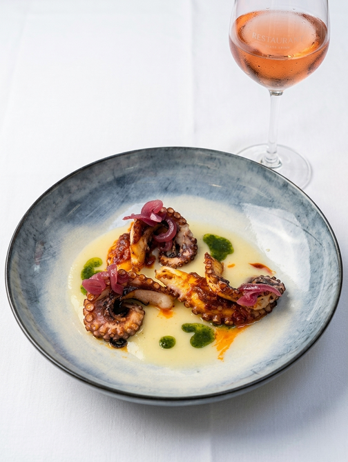 Piatto di polpo in una ciotola grigia con salsa verde, accanto a un bicchiere di rosé su un tavolo bianco.