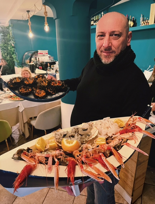 Un uomo regge un grande piatto di frutti di mare in un ristorante color turchese acceso, con altri piatti sul tavolo alle sue spalle.
