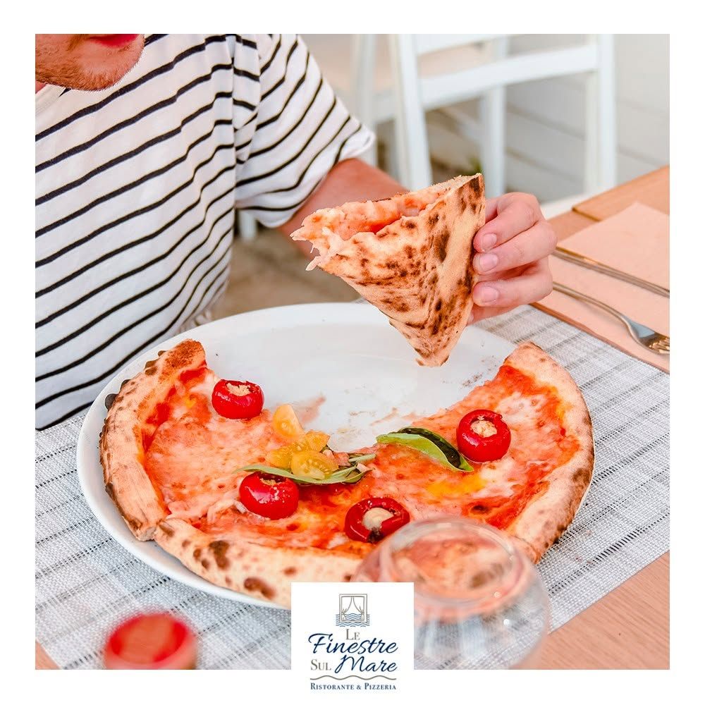 Una persona mangia una pizza con pomodorini a un tavolo, tenendo una fetta sopra un piatto bianco.
