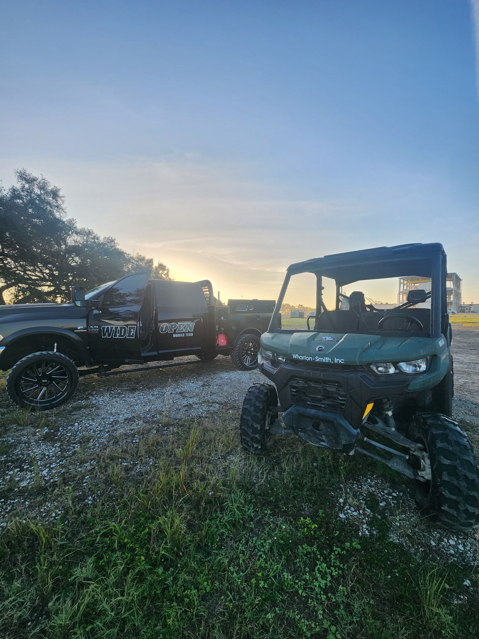 A dark pickup truck and a green side-by-side vehicle parked on a grassy field at sunrise.