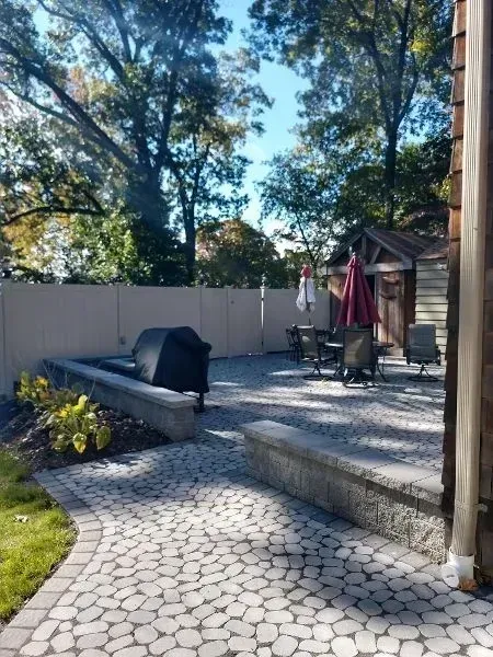 Patio with gray stone pavers, stone walls, grill, and outdoor seating. Trees and sky in background.