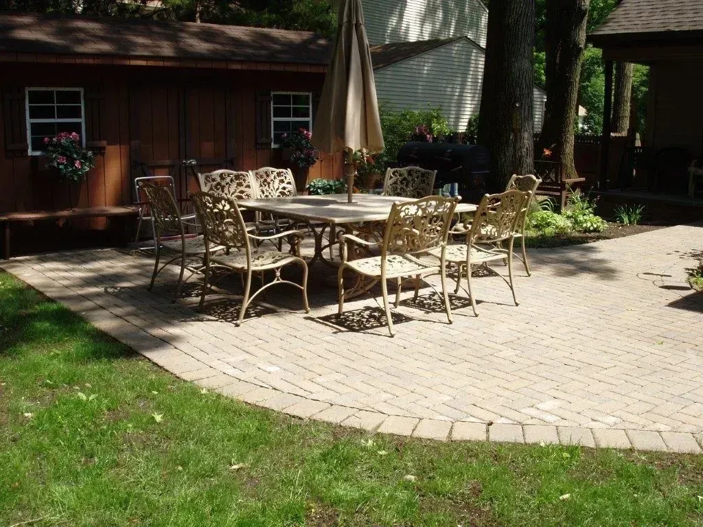 Outdoor patio with table, chairs, and umbrella on brick pavers, beside a shed and grass.
