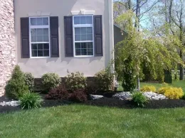 House exterior with windows, shutters, and a landscaped bed of shrubs, mulch, and river rock.