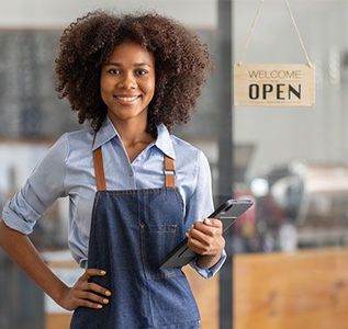 A woman in an apron is holding a tablet in front of a welcome open sign.