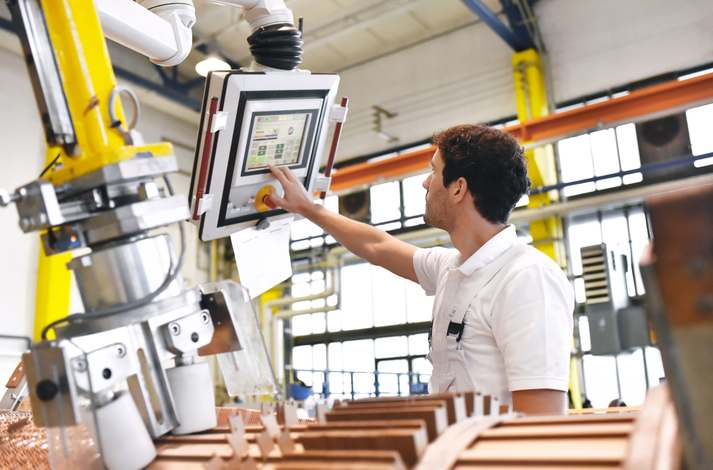 A worker in a factory adjusts settings on a control panel attached to a piece of robotic industrial equipment.