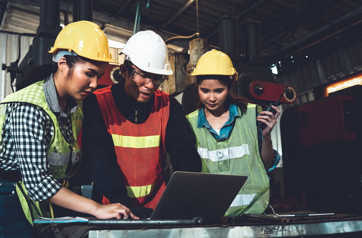Three factory workers wearing high-visibility vests and hard hats look at a laptop screen in an industrial setting.