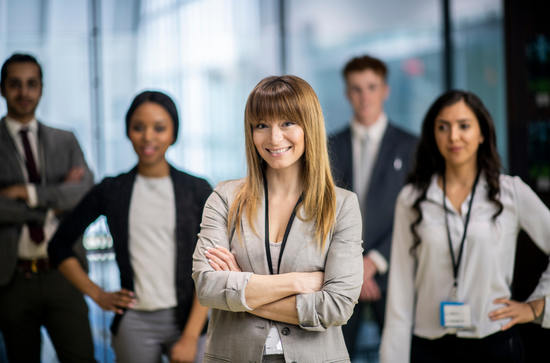 A group of five professionals in business attire stand in an office setting, with one person in focus in the foreground.