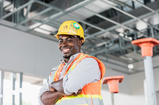 A smiling construction worker wearing a yellow hard hat and orange safety vest, standing with arms crossed on a job site.