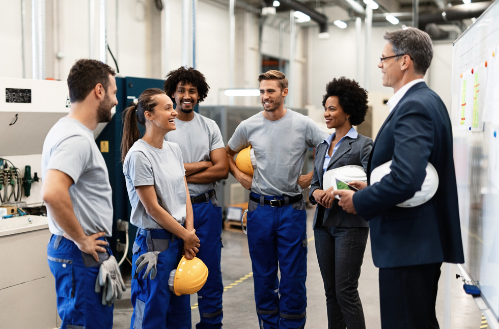 A supervisor in a suit speaks with a diverse group of industrial workers in uniform inside a factory setting.