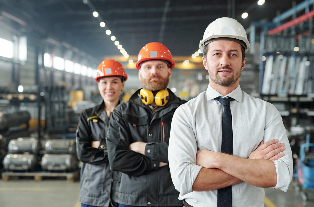 Three professionals in hard hats and work gear stand with arms crossed in a factory setting.